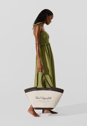 Suur koorekastme käekott tumepruunide detailidega ja musta värviga brändi nimi trükitud. Mudel kannab rohelist kleiti smock-kudumisega rinnaosas.