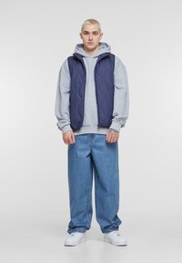 Navy quilted vest over a gray hoodie, paired with loose blue denim jeans and white sneakers. Smooth texture, relaxed fit, minimal design.