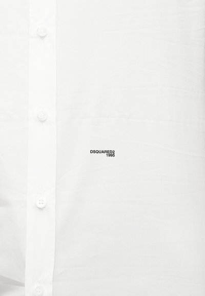 Chemise blanche à bouton avec un petit logo noir "DSQUARED2 1995" imprimé près de la poitrine, boutons visibles le long de la patte de boutonnage avant.