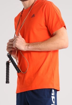 Persoon die een oranje Adidas T-shirt en marineblauwe shorts draagt, met een springtouw met zwarte handvatten om de nek.