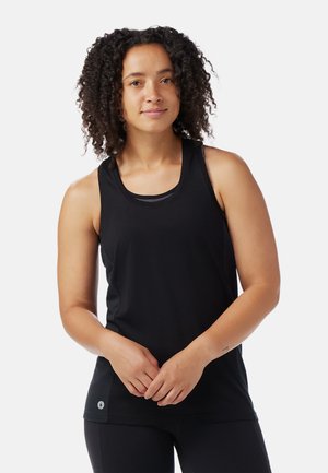 Donna con capelli ricci che indossa un top sportivo nero senza maniche e leggings neri, in piedi contro uno sfondo bianco semplice.