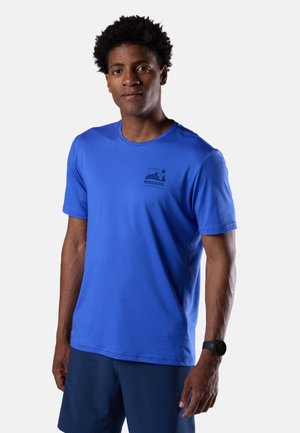 Uomo con capelli corti e ricci, indossa una T-shirt blu Rossignol e pantaloncini blu scuro, in piedi davanti a uno sfondo chiaro e uniforme.