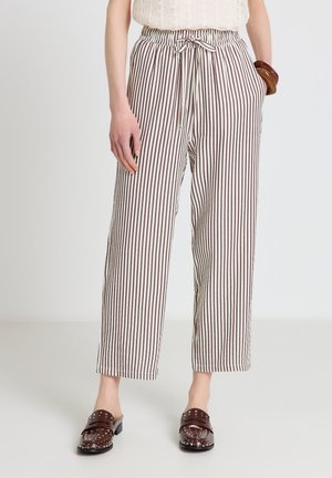 Donna che indossa pantaloni bianchi a vita alta con righe verticali marroni, vita con coulisse e scarpe slip-on marroni borchiate, in piedi con le mani nelle tasche.