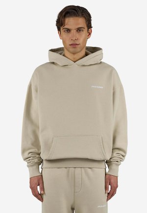Ung mand iført en beige hættetrøje og matchende sweatpants med lille hvid logo tekst på brystet og låret.