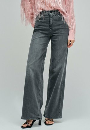 Femme portant un jean gris taille haute à jambes larges, des sandales à talons noires à brides, et un haut à manches longues rose avec une texture plumeuse.