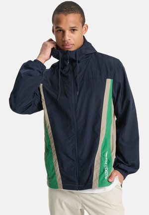 Navy windbreaker jacket featuring green and beige diagonal stripes, adjustable hood, elastic cuffs, and front zipper. Lightweight fabric.