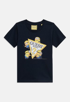 Teget plava majica s četiri Miniona koji stupaju u interakciju s velikim žutim trokutastim logom na kojem piše "GUESS Minions."