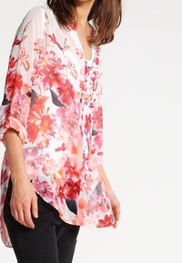 Blusa floreale con motivi di fiori rosa e rossi su un tessuto traslucido, con chiusura a bottoni e maniche arrotolate.
