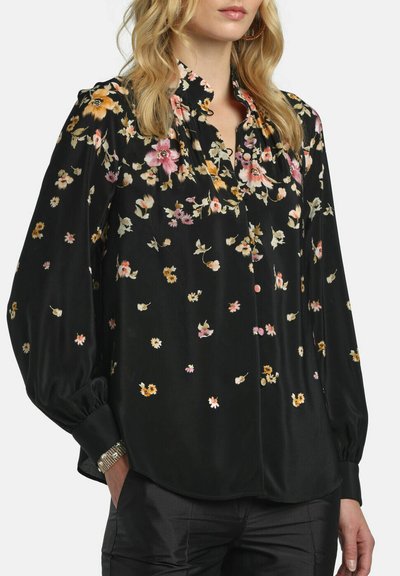 Femme portant un chemisier noir à manches longues avec des motifs floraux dispersés en rose, orange et blanc, associé à un pantalon noir et une montre argentée.