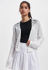 Weißer Cardigan mit einem zarten Spitzeneinsatz an den Schultern, kombiniert mit einem schwarzen Top und einem gefalteten weißen Rock. Leicht, langärmlig.