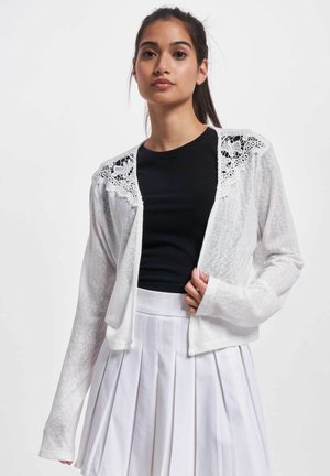 Weißer Cardigan mit einem zarten Spitzeneinsatz an den Schultern, kombiniert mit einem schwarzen Top und einem gefalteten weißen Rock. Leicht, langärmlig.