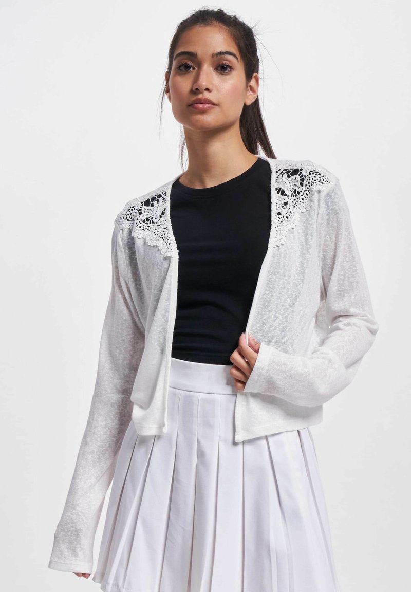 Weißer Cardigan mit einem zarten Spitzeneinsatz an den Schultern, kombiniert mit einem schwarzen Top und einem gefalteten weißen Rock. Leicht, langärmlig.