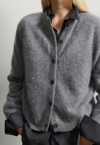 Grauer Wollcardigan mit weicher Textur, ausgestattet mit einer knöpfbaren Front, rundem Ausschnitt und leicht übergroßem Schnitt. Schwarze Hemdmanschetten sichtbar.