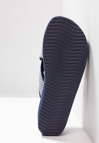 Chaussure avec une semelle en caoutchouc bleu marine foncé, présentant un motif ondulé texturé. Le dessus est conçu avec des lanières en tissu rayé bleu et blanc.