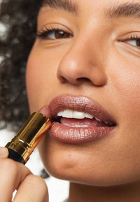 Rouge à lèvres dans un étui doré et noir appliqué sur des lèvres pleines et brillantes avec une teinte nude irisée. La peau est lisse avec un maquillage naturel.