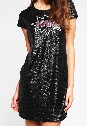 Femme portant une robe courte noire à sequins avec des manches courtes et le texte rose "KAR" à l'intérieur d'un motif éclat blanc sur la poitrine.