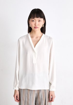 Chemisier blanc à manches longues avec un col à patte et des poignets élastiqués, fabriqué en tissu léger. Associé à un pantalon rayé.