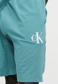 Ανοιχτό τιρκουάζ σορτς με λευκό λογότυπο Calvin Klein "cK" στο μηρό, φορεμένα από άτομο με σκουρόχρωμο δέρμα ορατό κάτω από το τελείωμα.