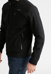 Veste softshell noire avec poches zippées, présentant un col haut et une finition texturée. Comprend un logo sur la poitrine et les coutures latérales.