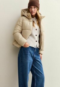 Vrouw draagt een beige pufferjack, een crèmekleurige gebreide cardigan, blauwe wijde jeans en een bruine gebreide muts, terwijl ze tegen een effen muur staat.