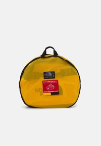 Žuta mrežasta torba s crnim detaljima i gornjom ručkom. Sadrži crvenu oznaku na kojoj piše "BASE CAMP DUFFEL L" i dimenzije.