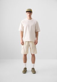 Light beige oversized t-shirt, cream shorts with side pockets, gray accents, white socks, and olive green sneakers. Mesh cap with logo.