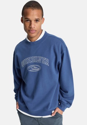 Junger Mann trägt einen blauen Quiksilver-Sweatshirt mit weißem Logo und posiert mit den Händen in den Taschen vor einem einfachen hellen Hintergrund.
