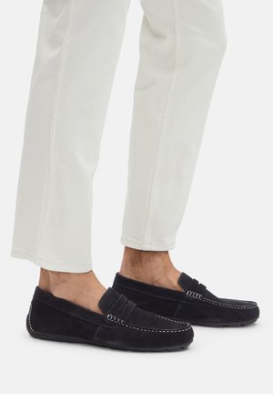 Zamszowe mokasyny w ciemnym granatowym kolorze, z płaską podeszwą, ozdobione dekoracyjnym przeszyciem i prostym paskiem. Noszone z białymi, krótkimi spodniami.