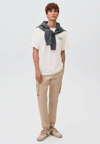 Ung mand iført beige cargo-bukser, hvid T-shirt med tekst, grå sweatshirt draperet over skuldrene og beige sneakers, stående imod en hvid baggrund.