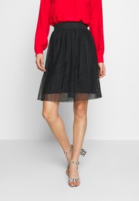 Schwarzer plissierter Rock mit tailliertem Bund, kombiniert mit Sandalen mit Zebra-Print und Absatz. Rotes, langärmeliges Oberteil, das am Bund sichtbar ist.