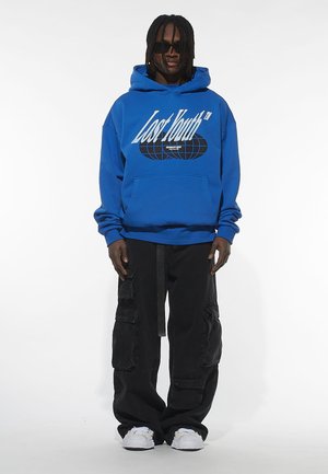 Blå hættejakke med "Lost Youth" grafik og globusdesign, parret med sorte cargo-bukser. Modellen bærer solbriller og hvide sneakers.