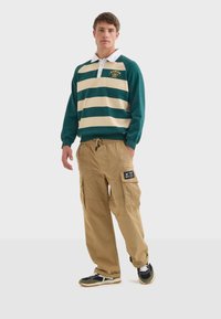 Zeleno-krémový pruhovaný polokošile s dlouhým rukávem a bílým límečkem, v kombinaci s béžovými cargo kalhotami s bočními kapsami a logem.