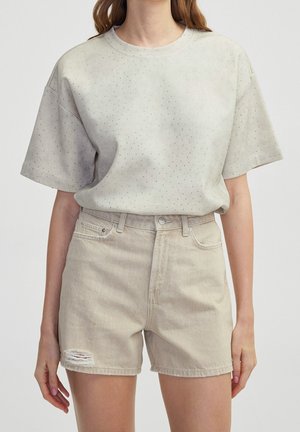 Kvinde iført en lysebeige prikket kortærmet skjorte og beige højtaljede denimshorts med en lille rift på venstre lår.