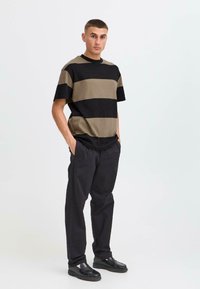 T-shirt à rayures vert olive et noir, manches courtes, coupe décontractée. Associé à un pantalon noir et des chaussures noires, avec une texture lisse.