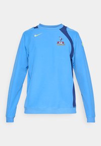 Gaiši zils džemperis ar garām piedurknēm, tumši zili akcenti uz pleciem, Nike logo dzeltenā krāsā un komandas emblēma priekšā. Mīksta auduma tekstūra.