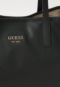 Svart texturerad tote-väska med dubbla handtag, prydd med guldlogotypen "GUESS EST 1981" på framsidan. Slät innerfoder synlig.