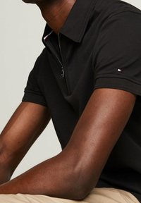 Svart polo skjorta med dragkedja vid kragen och korta ärmar. Har en liten logotyp på ärmen. Slät tygkvalitet med en åtsittande design.