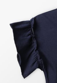 Námořnická modrá blůza s volánovými krátkými rukávy, vyrobená z měkké a pružné látky. Vyznačuje se hladkým povrchem a jednoduchým, čistým designem.