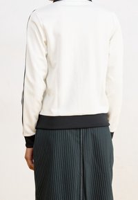 Felpa bianca con colletto e polsini neri, con strisce laterali nere a contrasto. Abbinata a una gonna verde scuro a righe sottili.