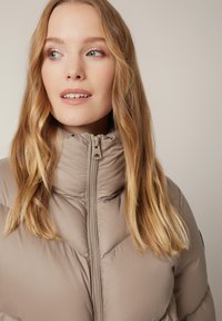 Blondynka z niebieskimi oczami spogląda w lewo, ubrana w jasnobeżową kurtkę puchową z zapięciem pod szyję, na jednolitym tle.