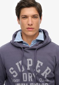 Sudadera morada con letras grises, material de algodón, capucha con cordón, usada sobre una camisa de cuello azul claro. Diseño simple y casual.