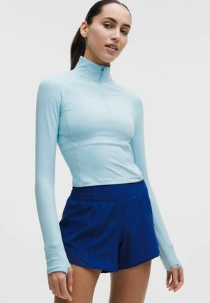 Kvinde iført lyseblå langærmet zip-up top og mørkeblå træningsshorts, stående mod en ensartet lys baggrund.