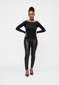 Μαύρο μακρυμάνικο εφαρμοστό τοπ συνδυασμένο με μαύρο δερμάτινο κολάν και σκούρα ψηλοτάκουνα παπούτσια· το μοντέλο στέκεται με τα χέρια στη μέση μπροστά σε λευκό φόντο.