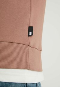 Body sweater in stofroze met een geribde zoom, voorzien van een zwart label met een witte vierkant, gedragen over een wit shirt.