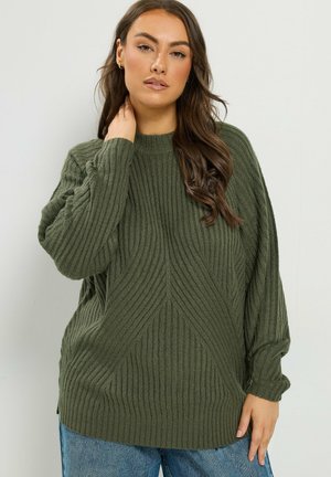 Femme aux longs cheveux bruns portant un pull en tricot côtelé vert et un jean bleu, touchant son cou d'une main.
