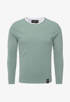 Pull à manches longues col rond vert atténué avec un bord blanc au niveau du col et une petite étiquette noire près de l’ourlet.