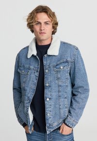 Giovane uomo che indossa una giacca di jeans blu con colletto in pile bianco sopra un maglione scuro, mani in tasca, su uno sfondo chiaro e semplice.