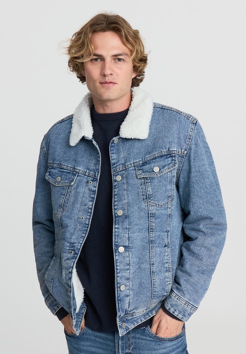 Giovane uomo che indossa una giacca di jeans blu con colletto in pile bianco sopra un maglione scuro, mani in tasca, su uno sfondo chiaro e semplice.