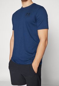 Muže ve modrém tričku s krátkým rukávem Under Armour a černých šortkách, s rukama v kapsách, stojí proti bílému pozadí.