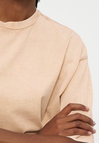 Άτομο που φοράει μπεζ κοντομάνικο πουκάμισο με σταυρωμένα χέρια, παρουσιάζοντας κοντινή λήψη του άνω κορμού και των χεριών.